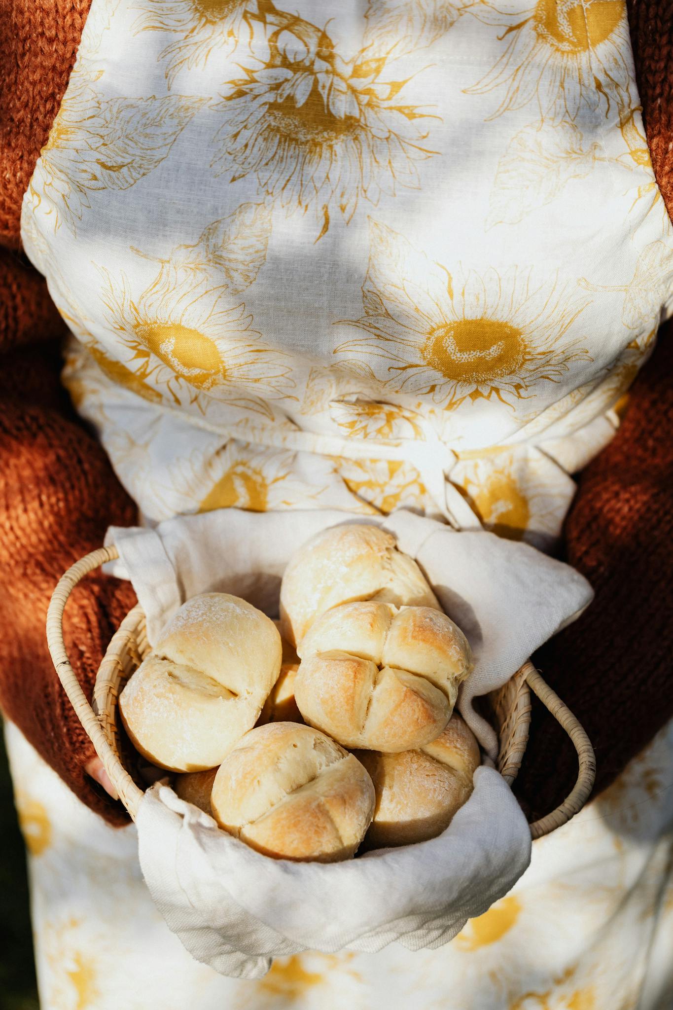Freshly baked buns in a woven basket, held by a person in a sunflower apron outdoors.